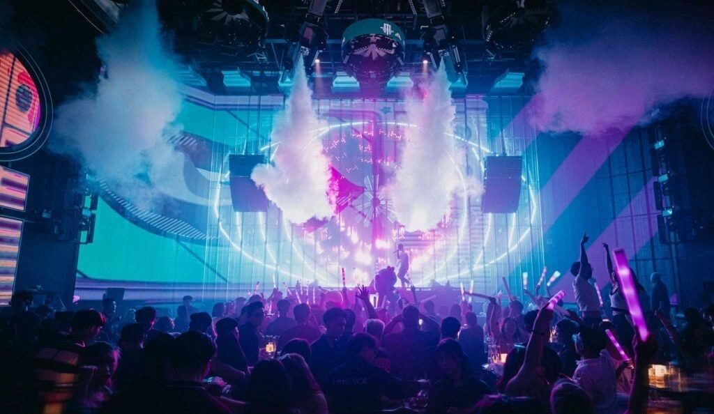 APLUS MUSIC SAIGON 舞台 CO2 炮喷射白雾，环形 LED 灯圈亮起，前排卡座坐满