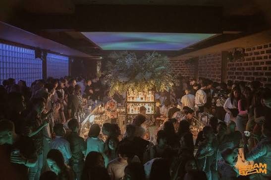 BAM BAR 高峰时段:吧台四周站满客人,顶部投下青绿色光,垂挂绿植装置在中央