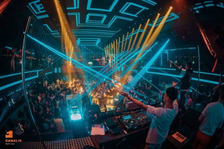 CANALIS CLUB DJ 台视角 · 橙蓝双色激光从两侧交叉射向舞池，DJ 举手带全场