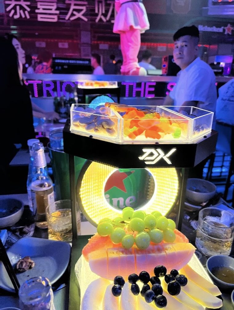 DISTRICT K 卡座上的发光果盘塔：底座嵌 DK 灯字与灯箱，上层三格亚克力盒装坚果软糖，背景是繁体霓虹招牌与满场客人