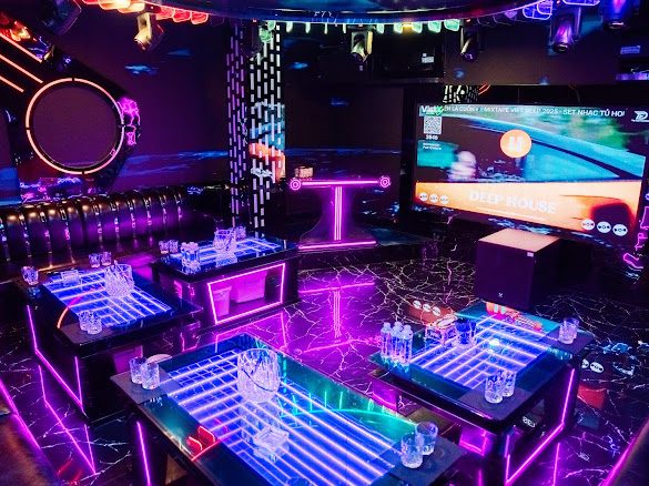 点歌大屏包厢:壁挂大屏正放越南语 Deep House 歌单,中间抬高的台阶用粉色灯带勾边,蓝紫发光茶几