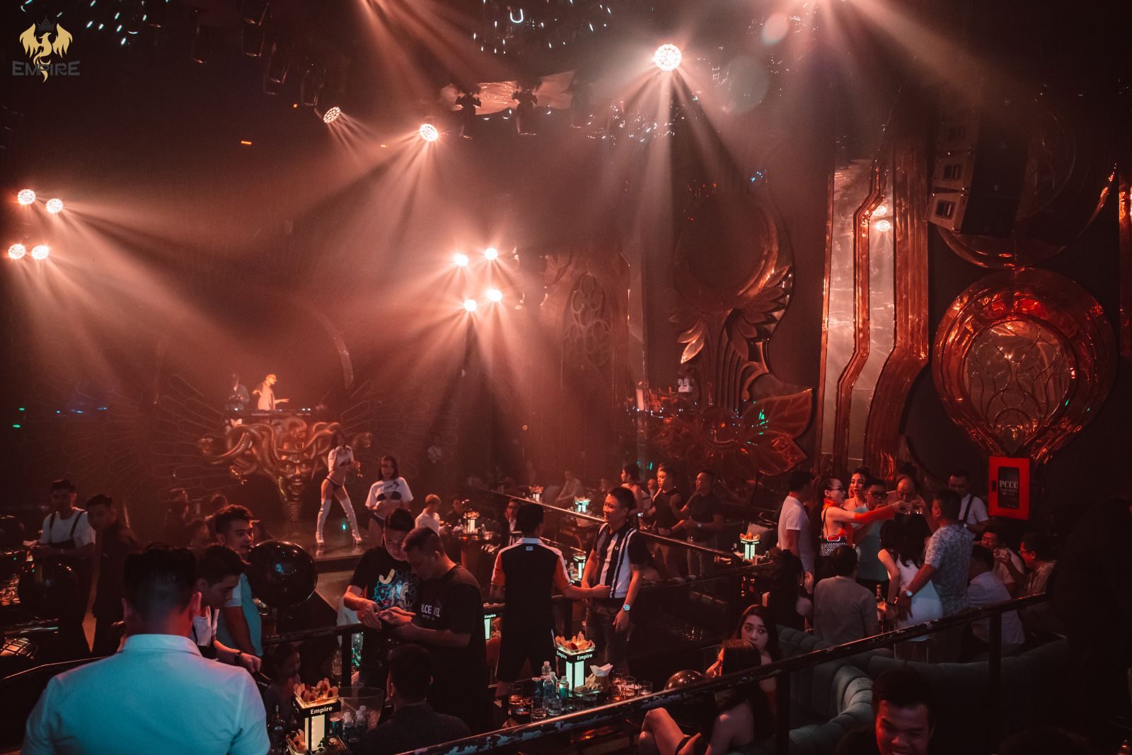 EMPIRE MUSIC BAR 胡志明第一郡音乐酒吧现场：铜色巴洛克浮雕墙面与美杜莎造型 DJ 台，舞者在中央舞台上，两侧阶梯卡座坐满客人，桌上摆着 Empire 字样的桌灯