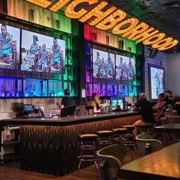 NEIGHBORHOOD BAR 彩色霓虹招牌墙与波普风画板，吧前排着绿色高脚凳