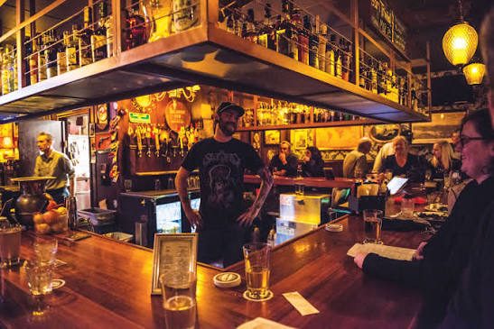 NEIGHBORHOOD BAR 暖木色长吧台，头顶悬挂式酒架，调酒师在吧内工作