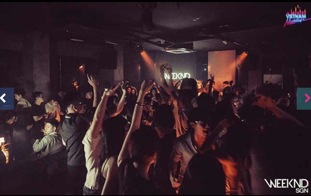 WEEKEND SAIGON 舞池全景，灯光压得很暗，人群举手朝向远处 WEEKND 字样的背墙