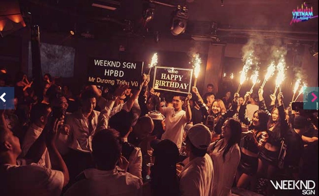 WEEKEND SAIGON 生日场面，两块 HAPPY BIRTHDAY 灯牌举在人群中，一排冷焰火同时点燃