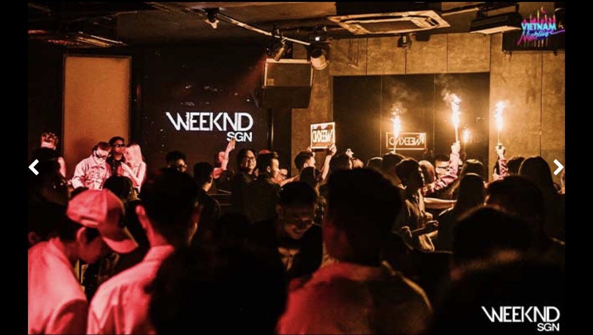 WEEKEND SAIGON 夜店现场，白色 WEEKND SGN 字样打在深色背墙上，客人举着冷焰火站满整个舞池